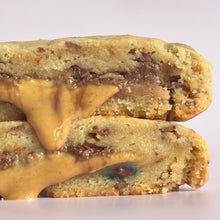 Load image into Gallery viewer, Two halves of a thick chocolate chip cookie stacked and shown close-up, with melted peanut butter sauce dripping over the cut surface and dark chocolate chips visible inside, on a plain light background.
